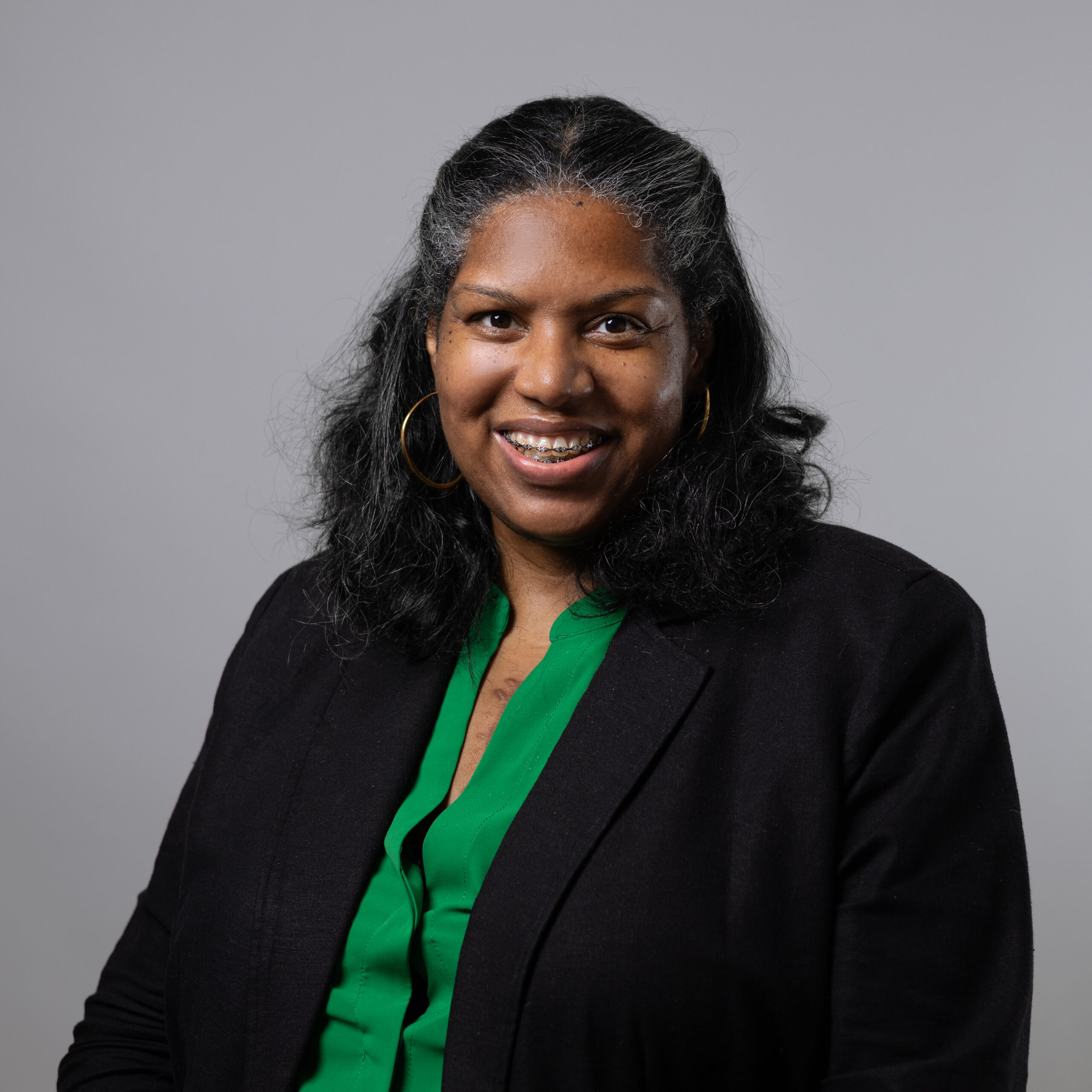 A smiling woman with long, curly black hair wearing large hoop earrings and a black blazer over a green blouse. The background is a neutral gray, and she appears friendly and confident.
