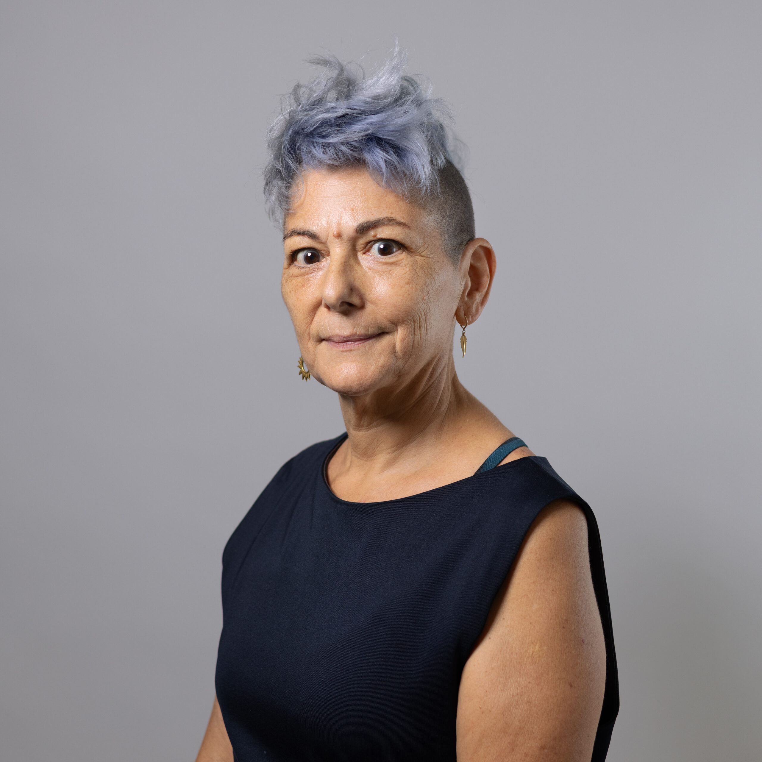 A woman with short, styled gray hair and a slight smile is looking at the camera. She is wearing a navy blue sleeveless top. The background is a light gray color. Earrings are visible in her ears, adding a touch of brightness to her appearance.