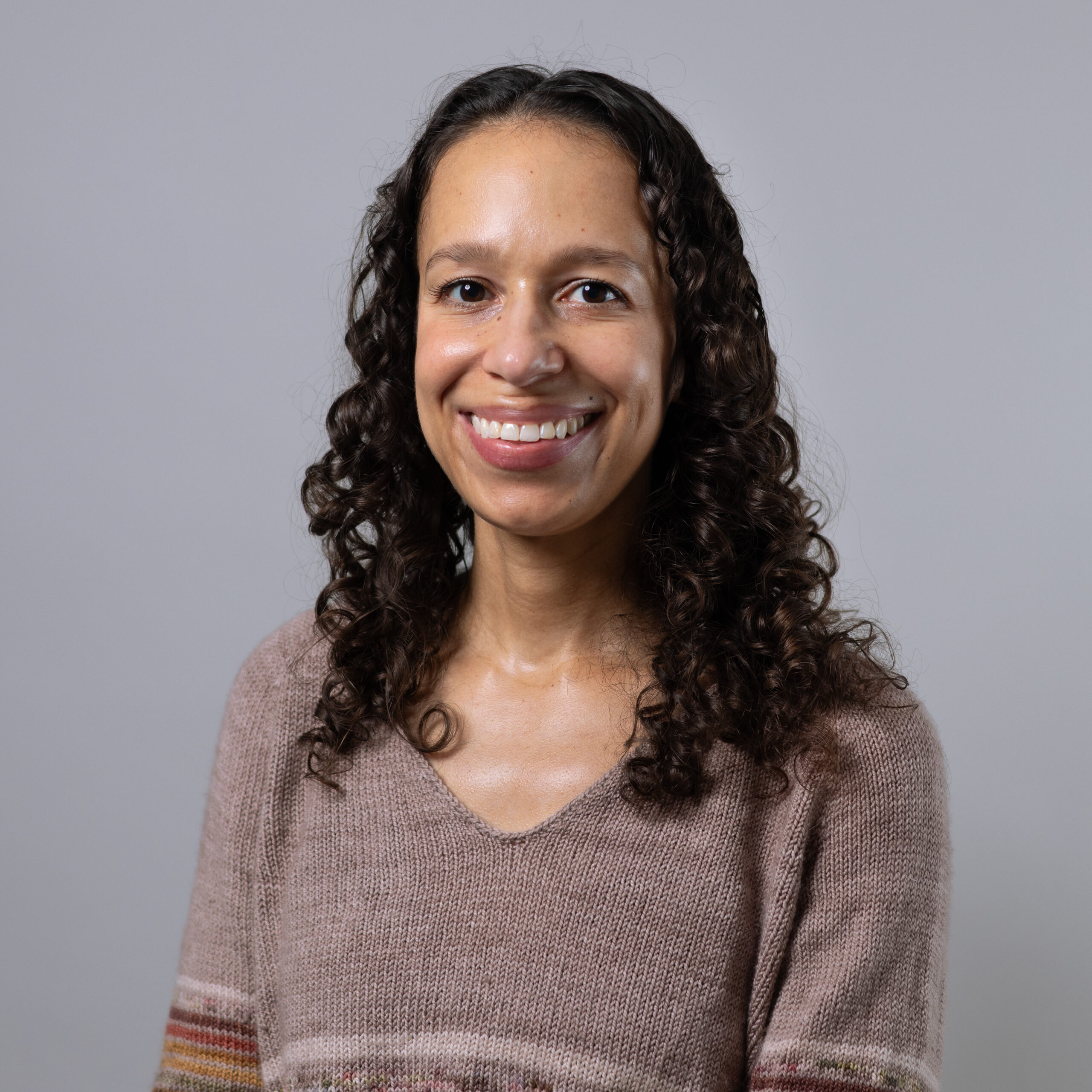 A smiling woman with curly hair is wearing a light brown sweater. The background is simple and neutral, emphasizing her cheerful expression.