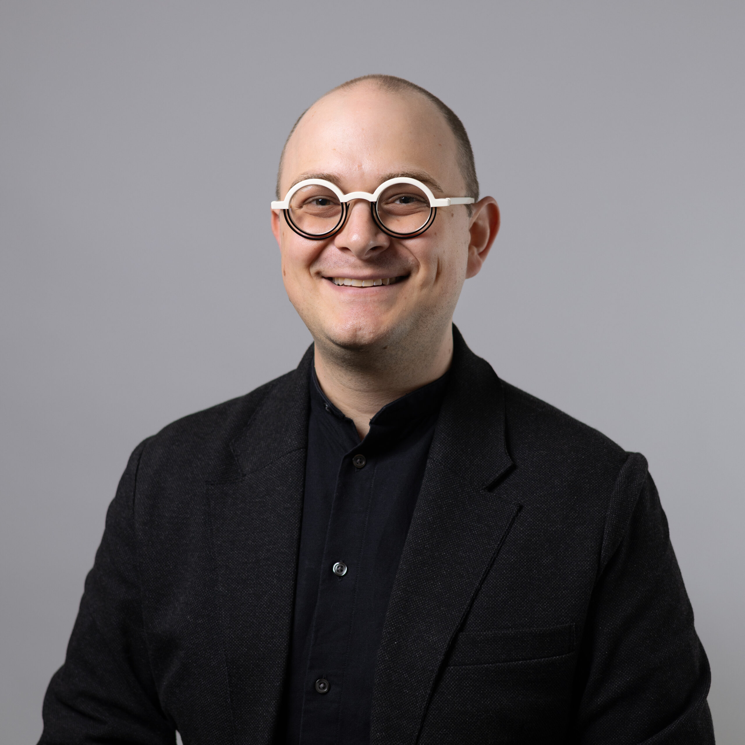 A smiling individual with a shaved head is wearing round, white-framed glasses and a dark jacket over a black shirt. They are positioned against a plain gray background, conveying a friendly and approachable demeanor.