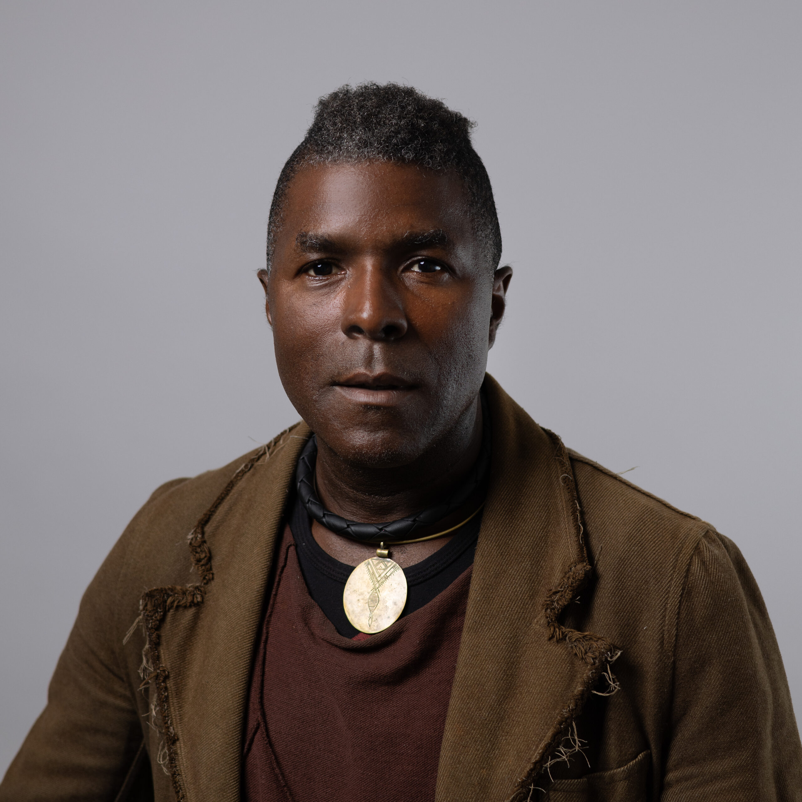 A close-up portrait of a man with a serious expression. He has short, styled hair with gray highlights and is wearing a layered outfit featuring a brown jacket with visible fraying, a dark top, and a necklace with a large circular pendant. The background is a simple gray, emphasizing his features and attire.
