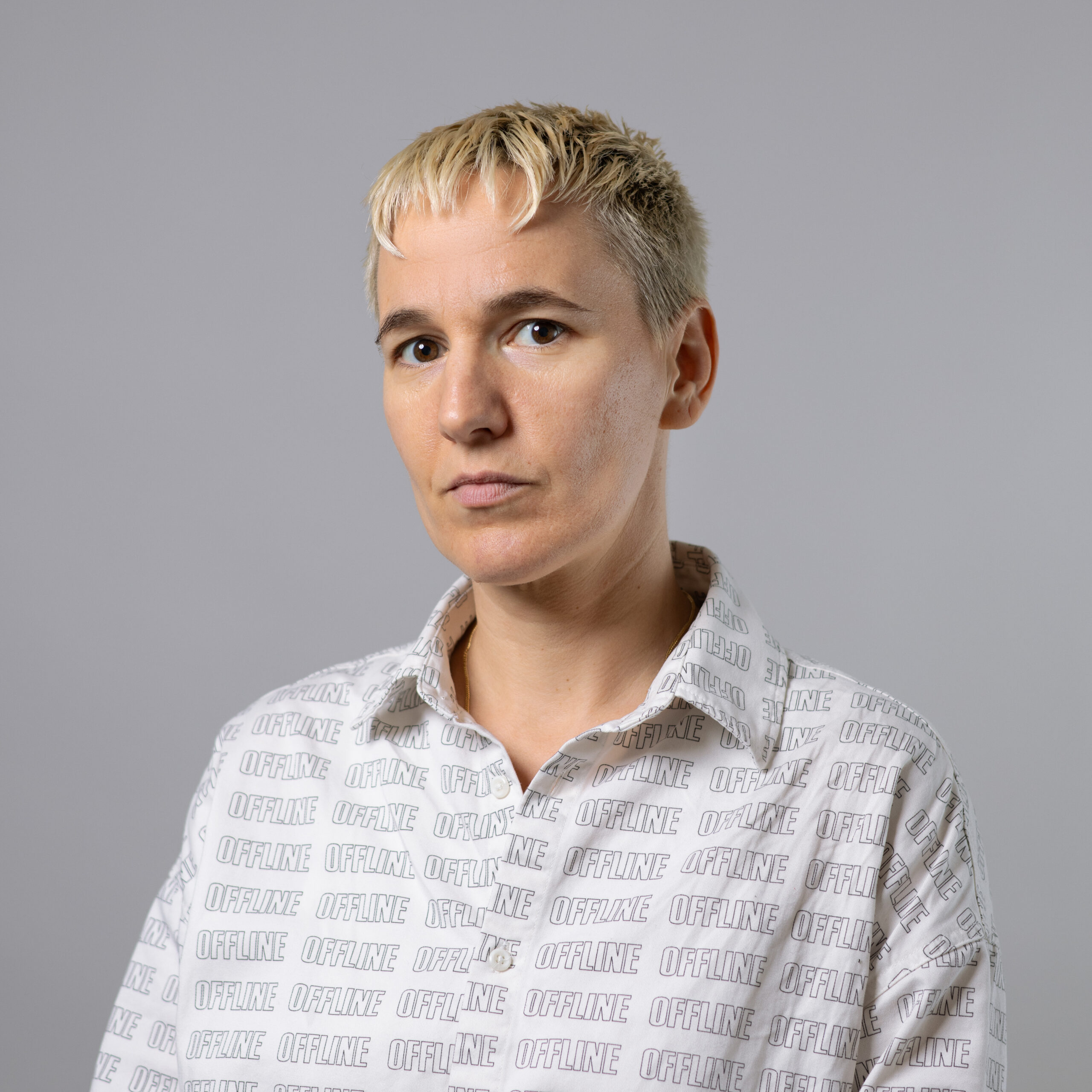 A person with short, light-colored hair is looking directly at the camera, wearing a white button-up shirt with the word "OFFLINE" printed in a repeating pattern. The background is gray, creating a neutral and focused atmosphere. The expression is serious and contemplative.