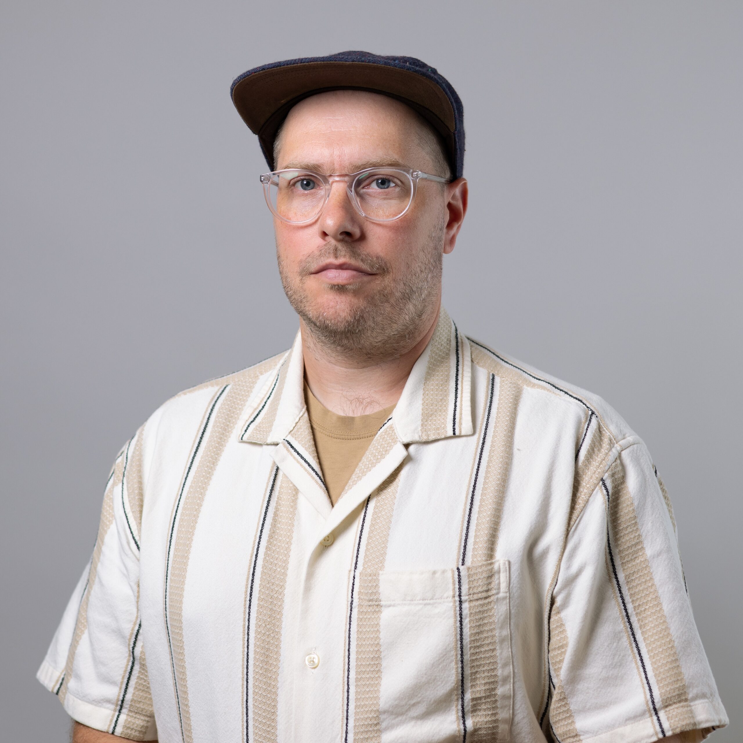 A person with short hair and glasses stands against a light gray background. They are wearing a patterned short-sleeve button-up shirt with horizontal stripes in cream and tan colors, layered over a plain tan t-shirt. A dark cap is worn at a slight angle. The expression is neutral, with a focus directed towards the camera.