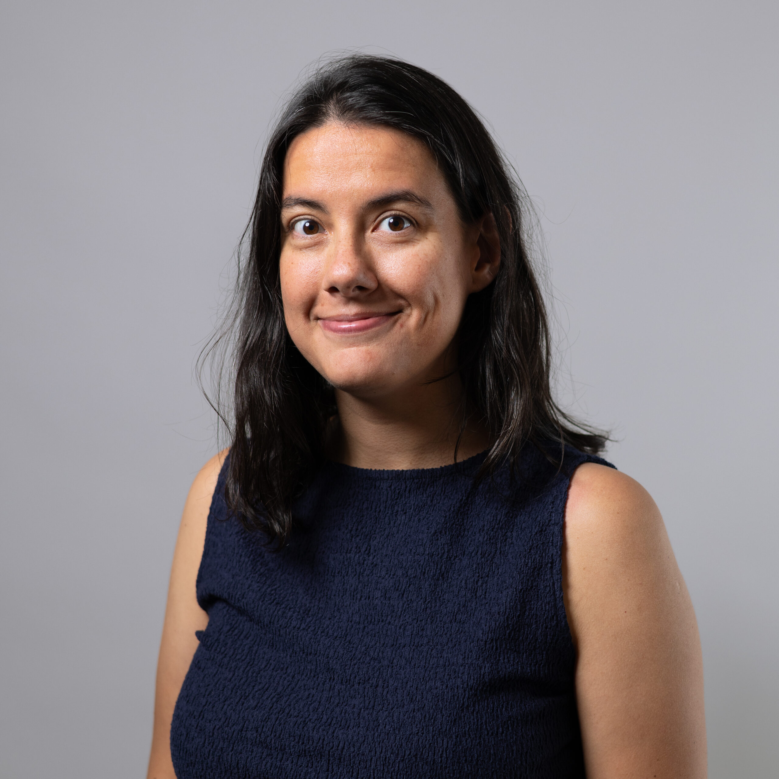 A smiling woman with long, dark hair is facing the camera. She is wearing a sleeveless navy blue top and has a friendly expression. The background is a light grey, providing a neutral contrast.
