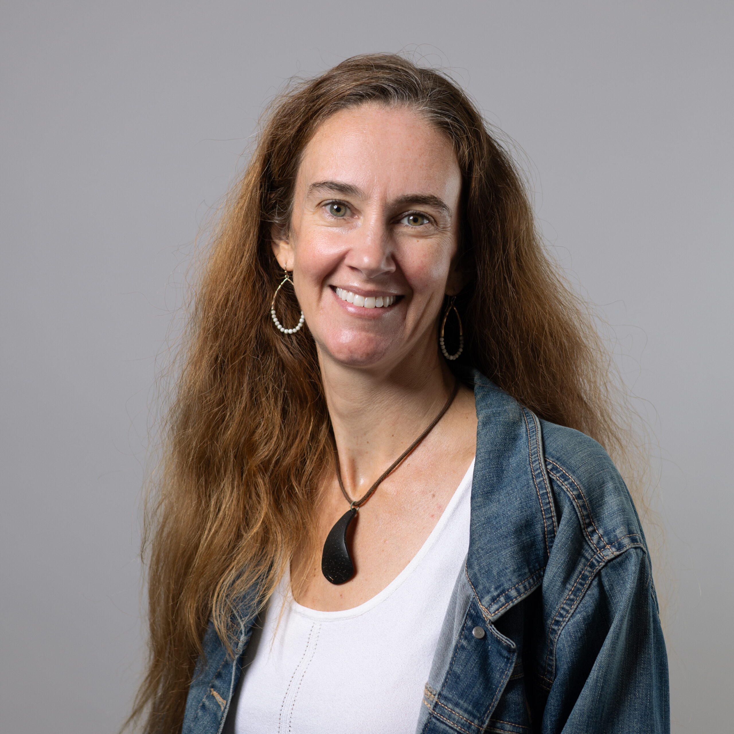 A woman with long, wavy brown hair smiles at the camera. She is wearing a denim jacket over a white shirt and a necklace with a black pendant. She has hoop earrings and is set against a light gray background.