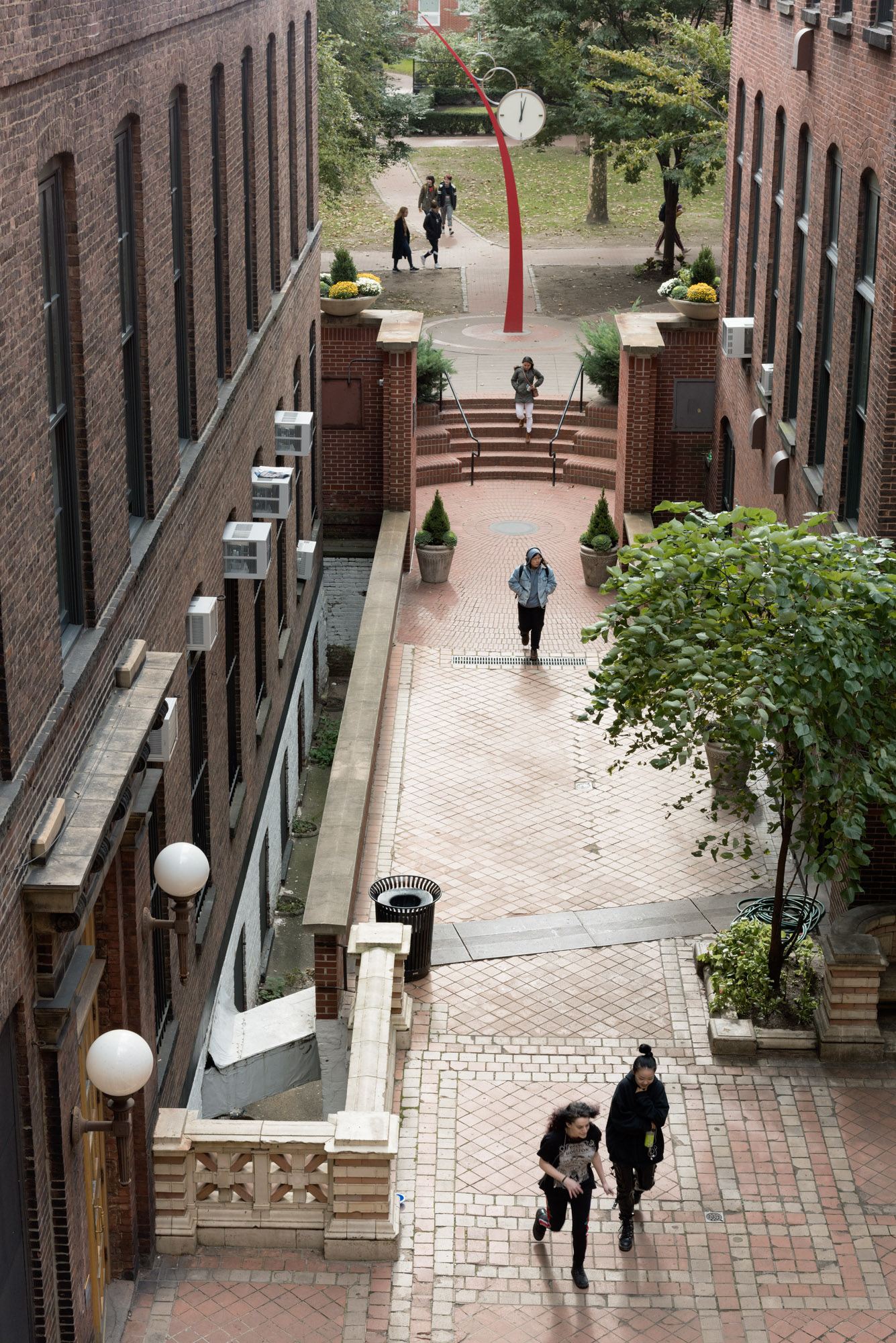 A view from above of a brick courtyard with several people walking along the pathways. In the foreground, two individuals are walking together. To the left, there is a set of stairs leading up to a platform. A striking red clock tower is visible in the background, surrounded by trees and flower pots. The area is paved, and there are benches and trash bins scattered throughout.