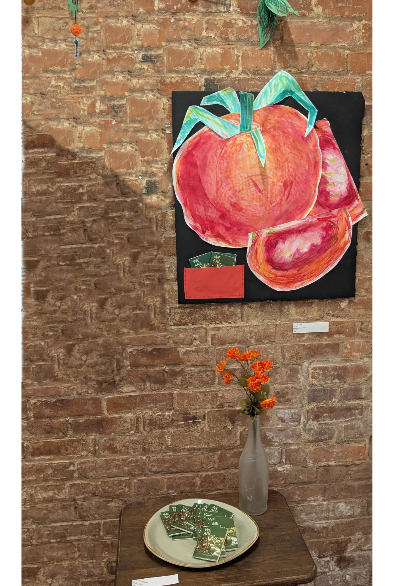 A colorful artwork depicting large, red fruits, possibly pomegranates, is mounted on a brick wall. Below the artwork, there is a small wooden table displaying a light green plate with several green pamphlets on it. A vase containing orange flowers sits beside the plate. A white label with text is visible near the art and table, and the background features rustic exposed brick.