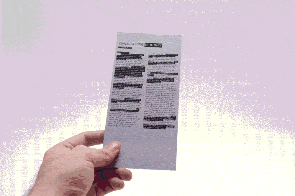 A person holding a light blue pamphlet titled "CONVERSATIONS IN BINARY" with text printed in black. The pamphlet includes various sections of text, some parts highlighted. The background appears blurred, focusing on the pamphlet and the person's hand.
