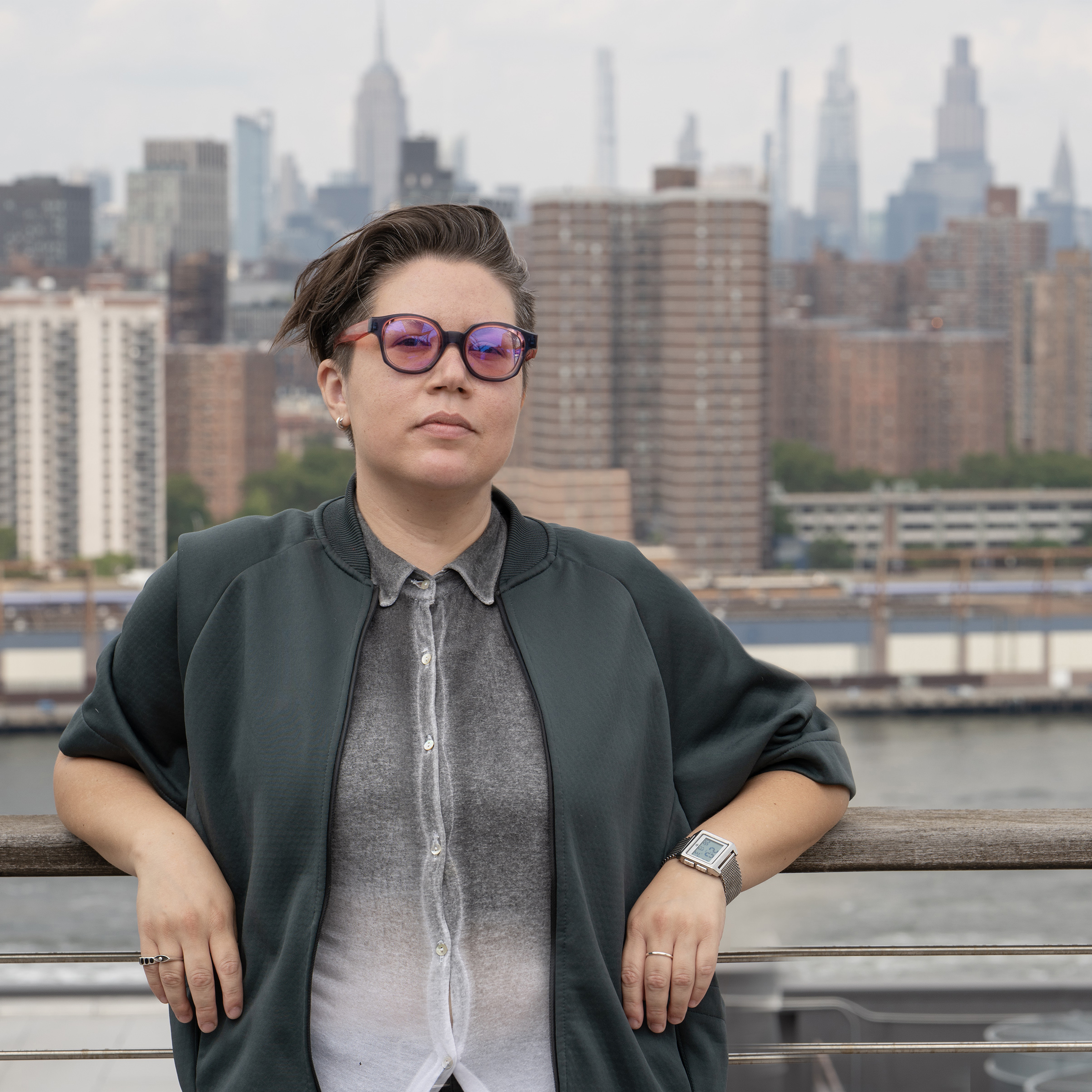 Photo of Mev Luna by Jesse Meredith. A person in glasses and a dark jacket stands by the waterfront with the New York City skyline in the background.