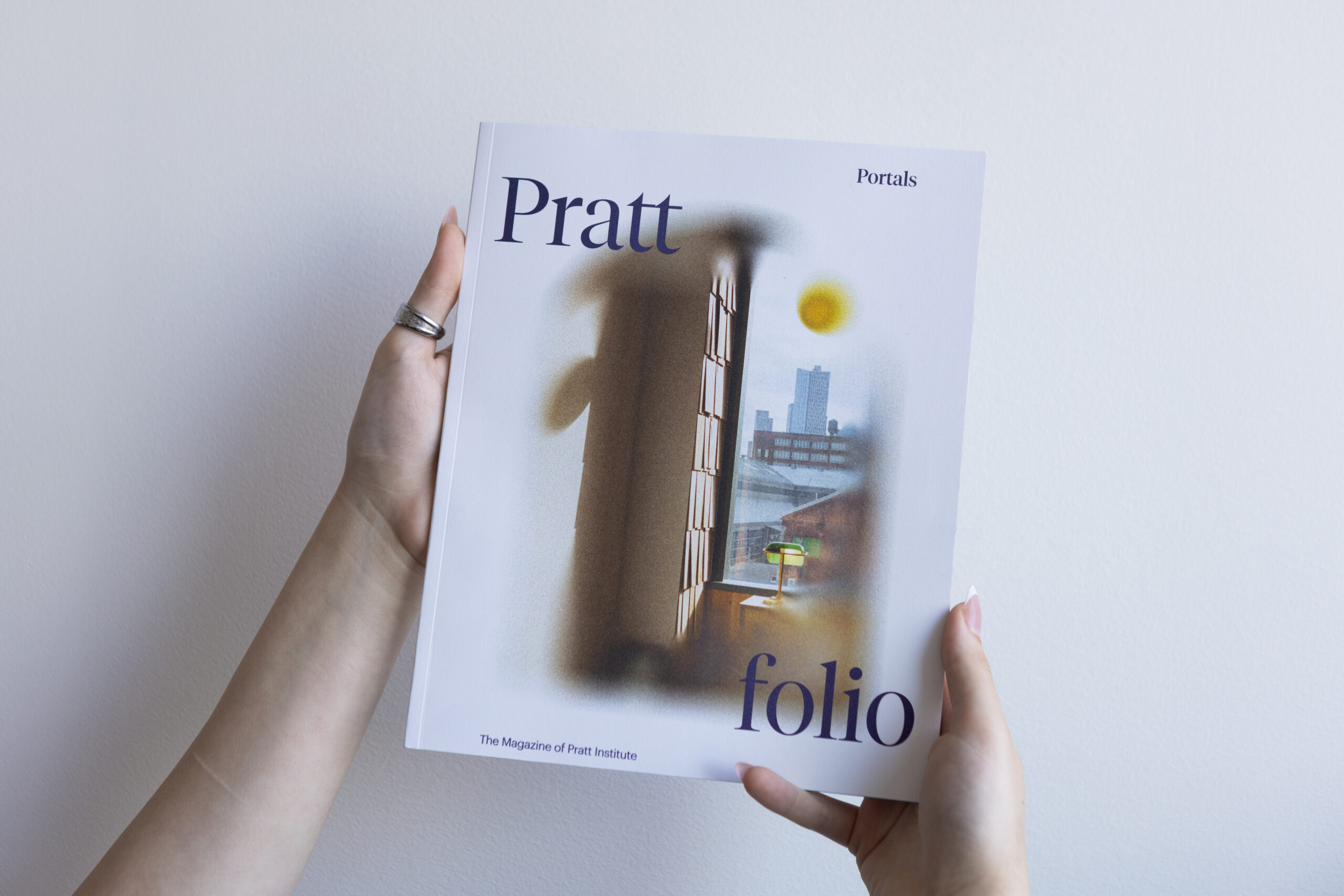 A reader holds up a copy of the spring 2026 issue of Prattfolio. The cover features a photograph of a the Brooklyn Navy Yard as seen from a studio window.