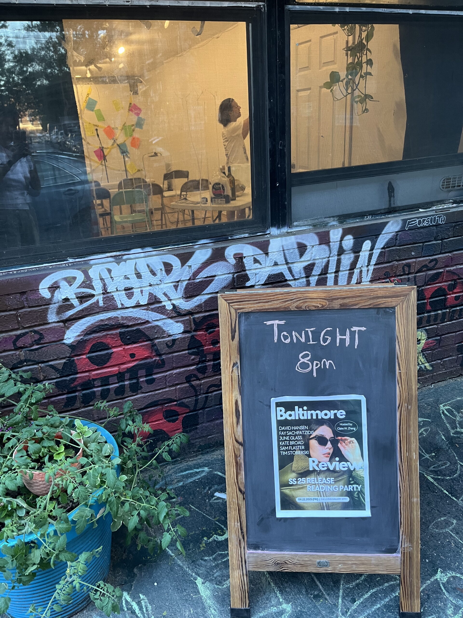 Outside of a brick-walled gallery with walls decorated with paintings of ladybugs, a sandwich board advertises the Baltimore Review spring/summer 25 release reading party. Inside the gallery, a person decorates the space.