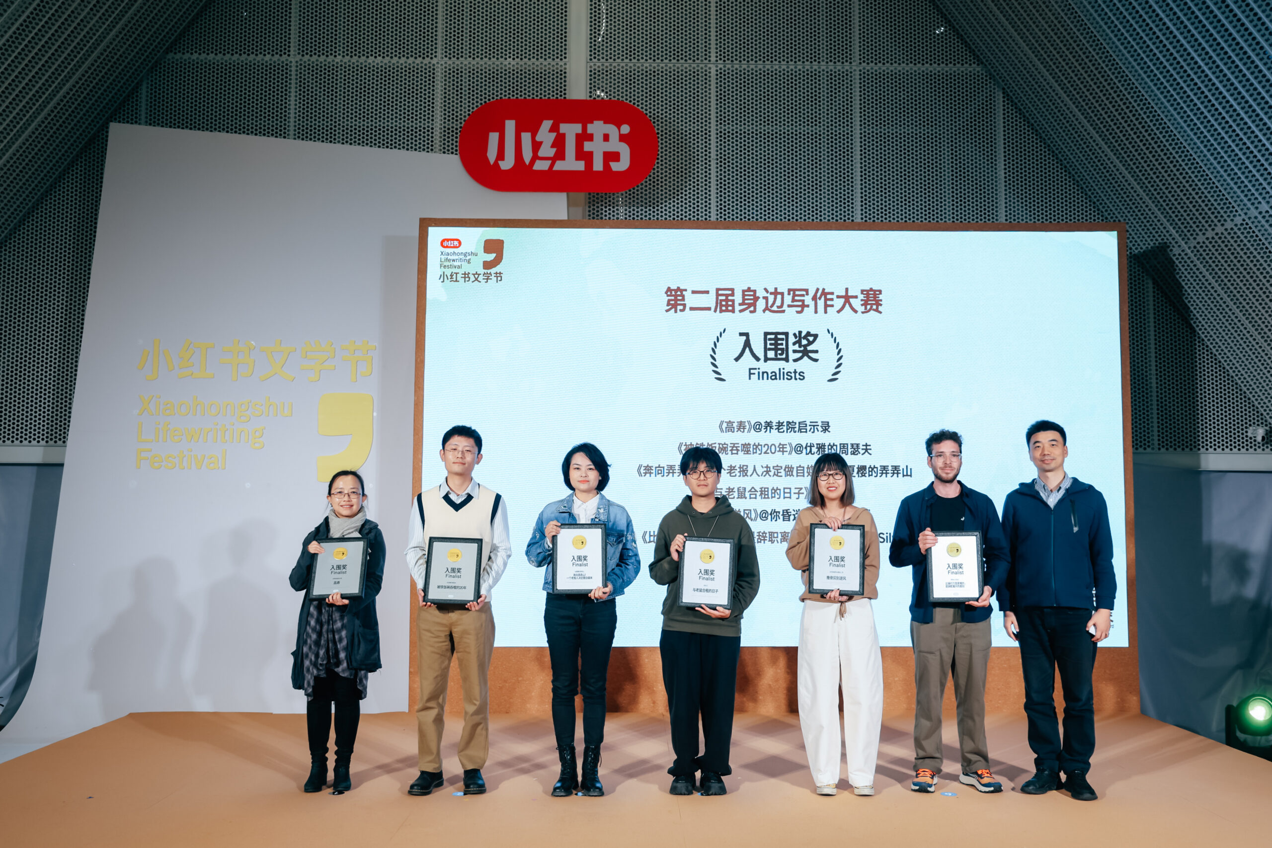 Alumna Claire W. Zhang stands on stage with other award recipients. They each hold an award. Signage reads, "Xiaohongshu Lifewriting Festival Finalists"