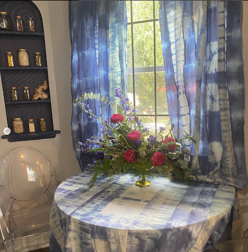 A cozy corner featuring a round table with a blue and white patterned tablecloth. On the table, there is an elegant floral arrangement with pink roses and greenery. Behind the table, sheer blue curtains frame a window that lets in natural light. A dark shelf on the wall displays jars, adding a decorative touch to the space. An acrylic chair sits beside the table, enhancing the inviting atmosphere.