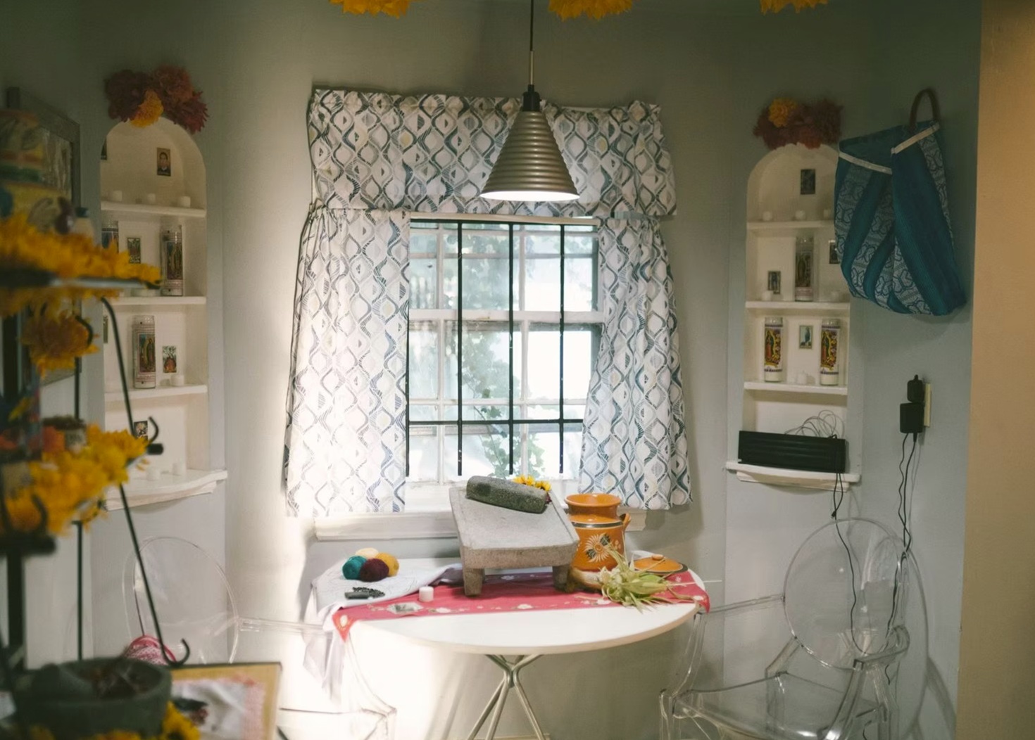 A cozy, well-lit room features a window with patterned curtains, a round table covered with a red cloth, and a traditional stone grinding tool on top. On the table are colorful yarns and an orange clay pot. White shelves display candles and small decorative items, while flowers adorn the edges. A clear acrylic chair adds a modern touch to the space, which has a warm and inviting atmosphere.
