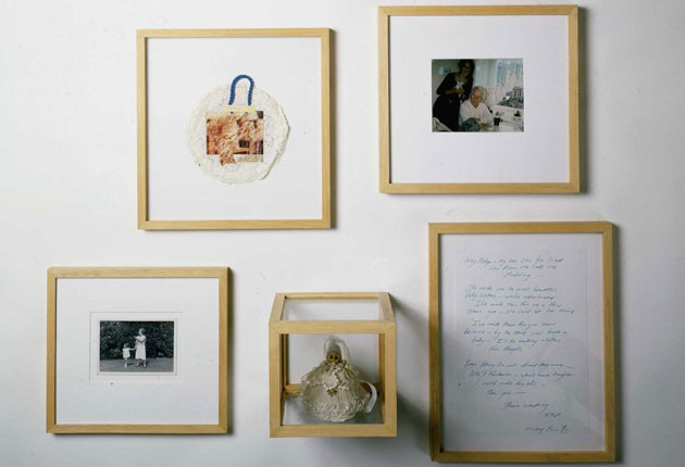 A collection of framed artworks arranged on a wall. The top left frame contains a textured circular piece with a blue handle and a photo. The top right shows a photo of a person at a table. The bottom left features an old black-and-white photograph of a child with a dog. In the center, there's a small display box containing a handmade doll. The right side holds a framed piece of handwritten text.