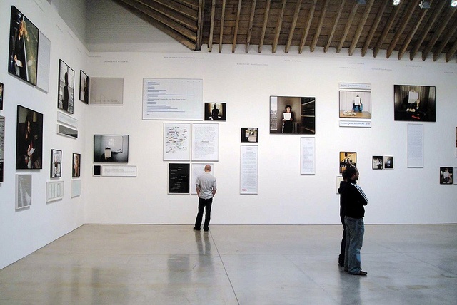 Two visitors examine a contemporary art exhibit featuring a collection of framed photographs and text arranged on a white wall. The interior space has a high, wooden-beamed ceiling, and the floor is smooth and polished. One person stands facing the wall, while another stands with their arms crossed, both engaged in observing the artwork displayed.