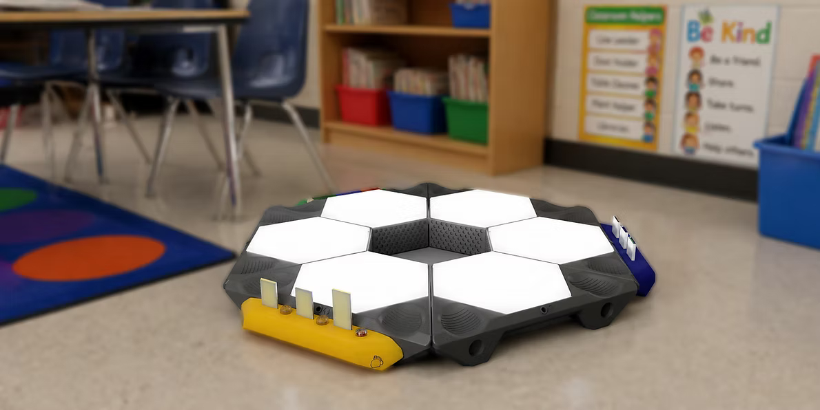 A hexagonal, colorful electronic device is placed on the floor of a classroom. It features multiple white sections on its surface and buttons in yellow and blue on the sides. In the background, there are bookshelves with bins and a wall displaying posters with educational messages. The floor has a multicolored circle mat in the corner.