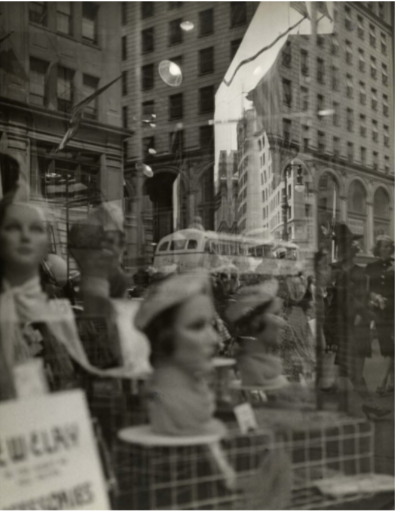 A black-and-white photo capturing a city street scene reflected in a store window. The foreground features several mannequins displaying hats, while the background reveals buildings and pedestrians. A bus and flags are visible in the reflection, contributing to a bustling urban atmosphere.
