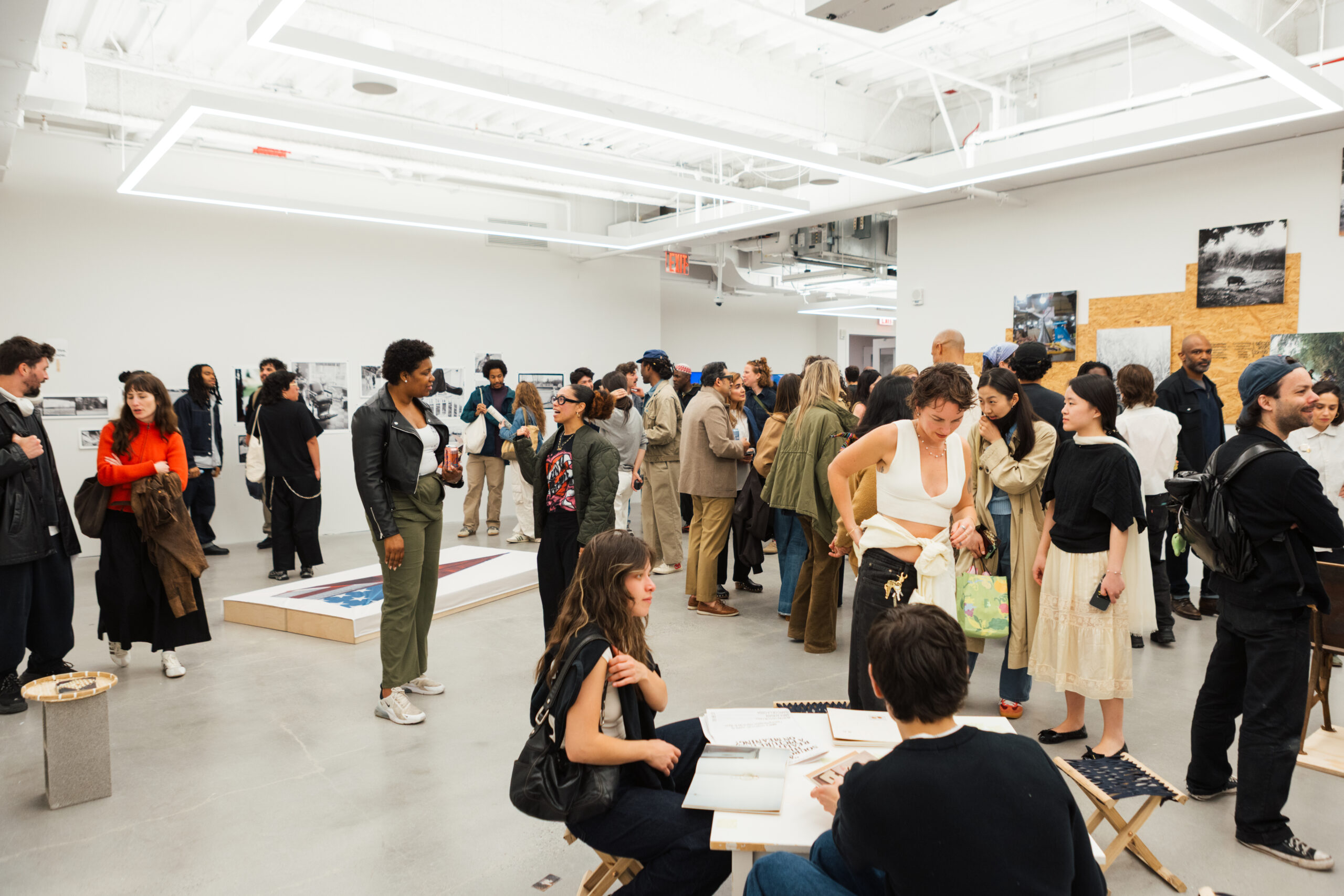 A crowded art gallery event featuring diverse attendees engaged in conversation. Groups of people are distributed throughout the space, some examining artworks on the walls while others socialize. Desks are visible with books and materials on them. The interior is bright with modern lighting and minimalistic decor.