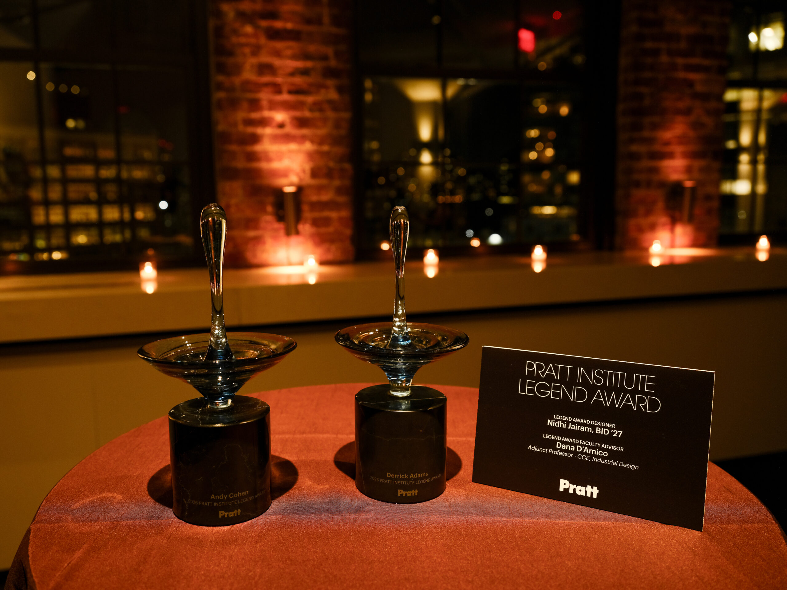 Two glass awards are displayed on a round table covered with a reddish-brown cloth. One award is inscribed with "Andy Cohen," and the other with "Derrick Adams." In front of the awards, a black card reads "Pratt Institute Legend Award" with details about the award designer and faculty advisor. Soft candlelight illuminates the background, creating a warm atmosphere.