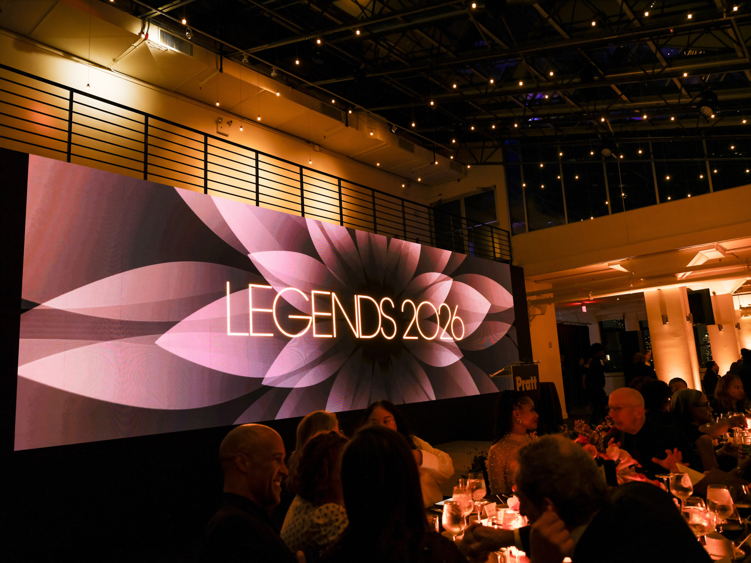 A large screen displays the text "LEGENDS 2026" against a soft, floral design with shades of pink. The setting is an elegant indoor event space with dim lighting and tables adorned with candles, where guests are seated, engaged in conversation. The ceiling features exposed beams and string lights, contributing to the festive atmosphere.