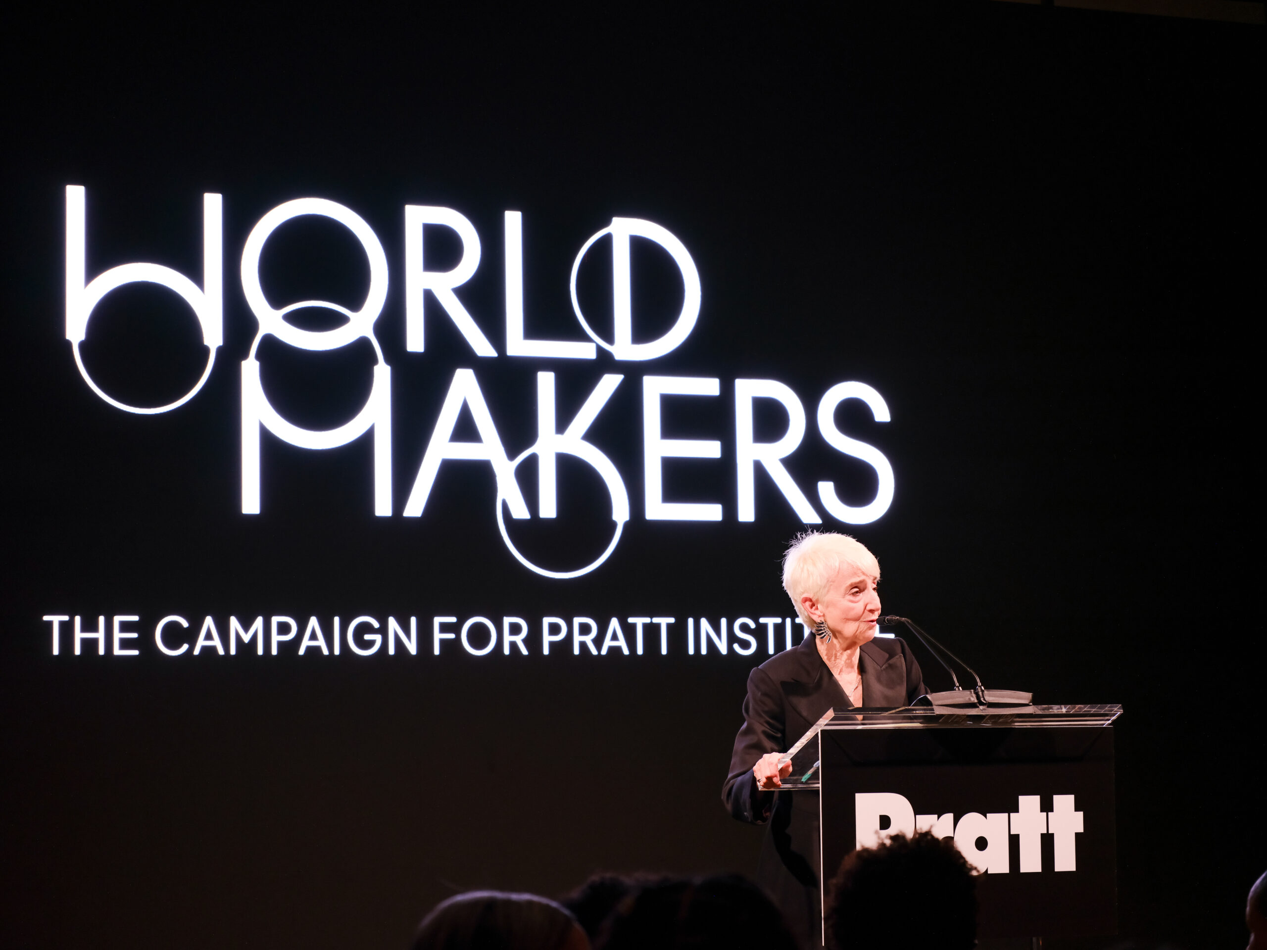A woman with short, white hair stands at a podium speaking during an event. The backdrop features the words "WORLD MAKERS" in large, stylish letters and a smaller text reading "THE CAMPAIGN FOR PRATT INSTITUTE." The setting appears formal and is focused on a presentation.