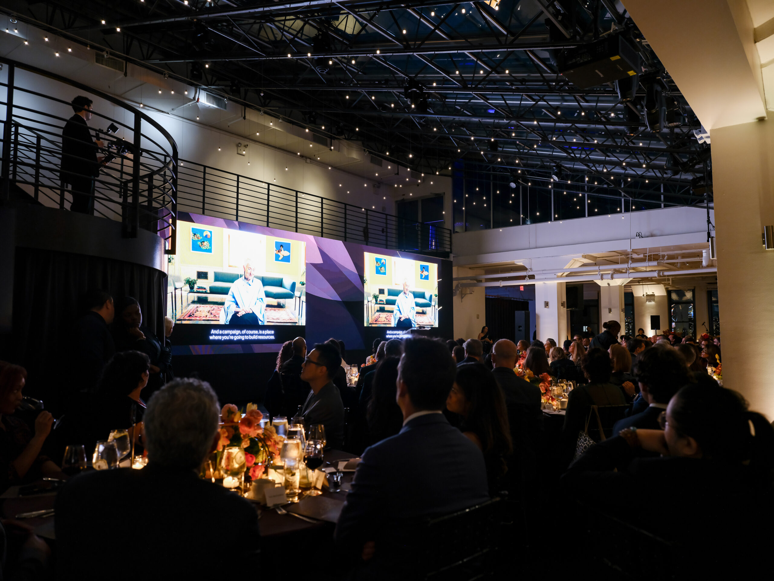 A formal event is taking place indoors, with attendees seated at elegantly set tables. In the foreground, a group of people is engaged in conversation, while a large screen displays a speaker talking about a topic, with subtitles visible. Decorative lighting hangs from the ceiling, adding to the ambiance. A person is seen on an elevated platform, possibly overseeing the event. The setting appears sophisticated and well-coordinated.