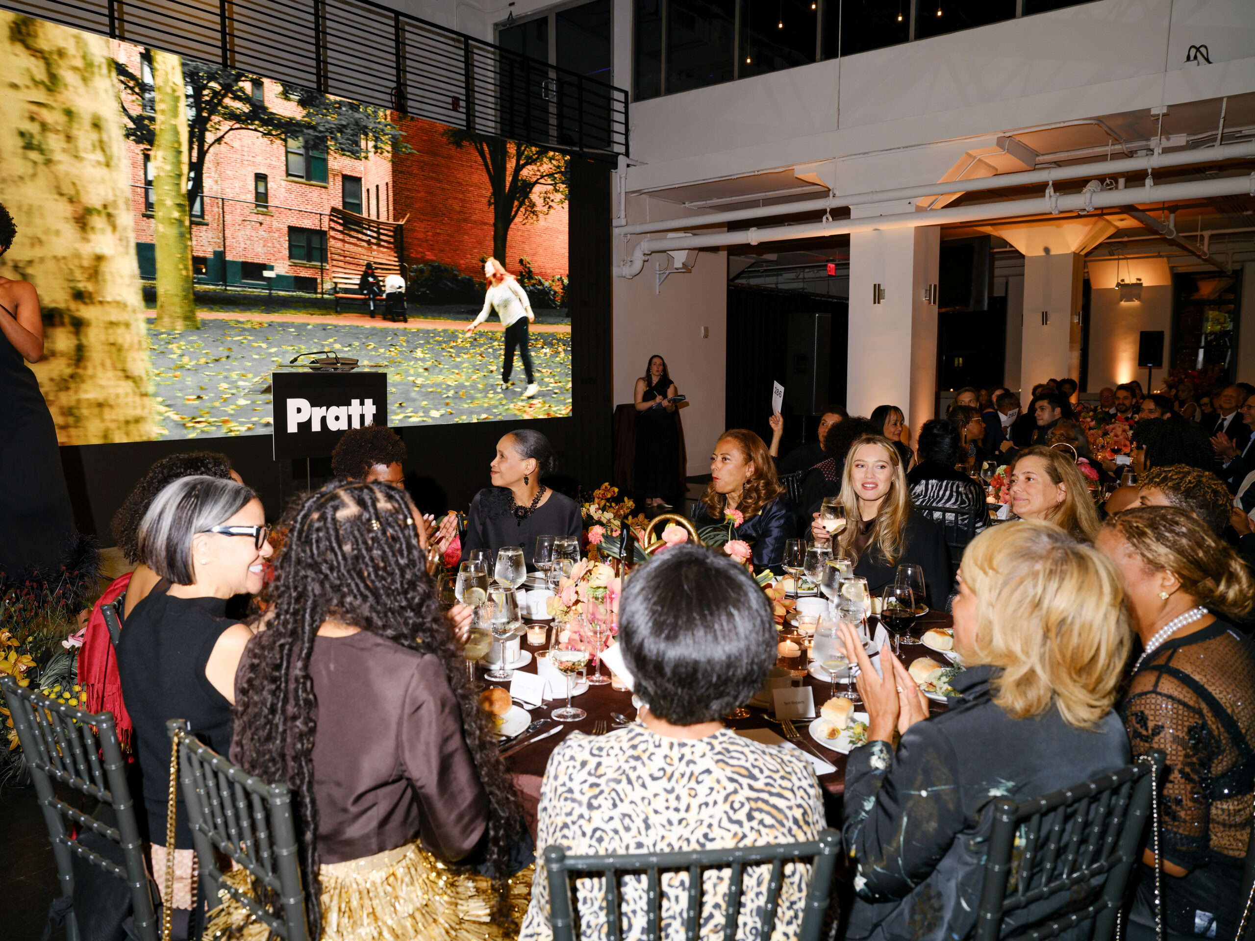 A group of elegantly dressed guests is seated around a decorated dining table during an event. In the background, a large screen displays a video of people in a park. The atmosphere is festive, with floral centerpieces and various glasses filled with beverages on the table. People are engaged in conversation, some smiling and clapping in response to the presentation.