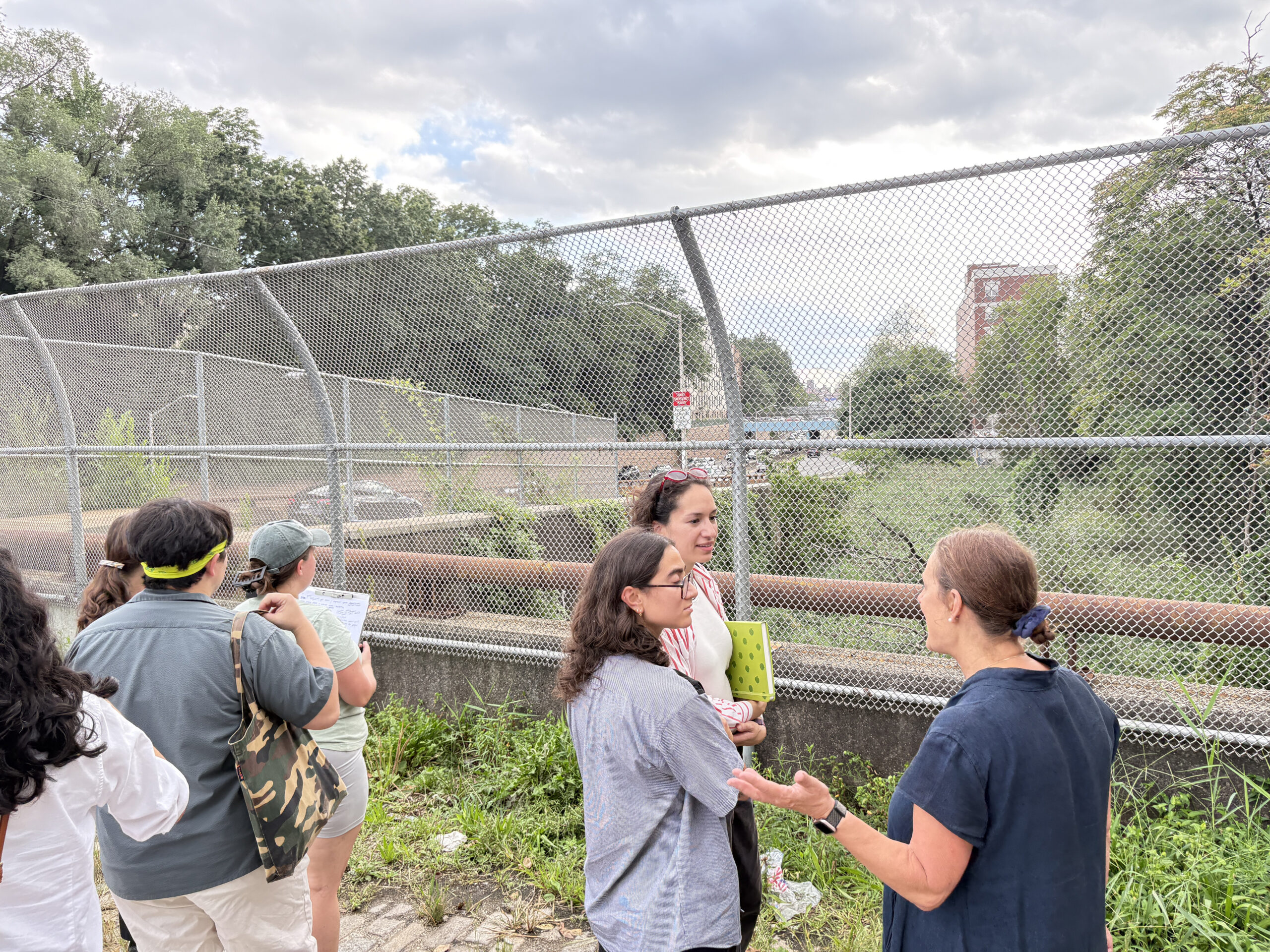 A group of four people stands by a chain-link fence overlooking a green area with trees and a road in the background. They are engaged in conversation, with one person holding a clipboard. The setting appears to be an urban environment, with buildings visible in the distance and overcast skies above.