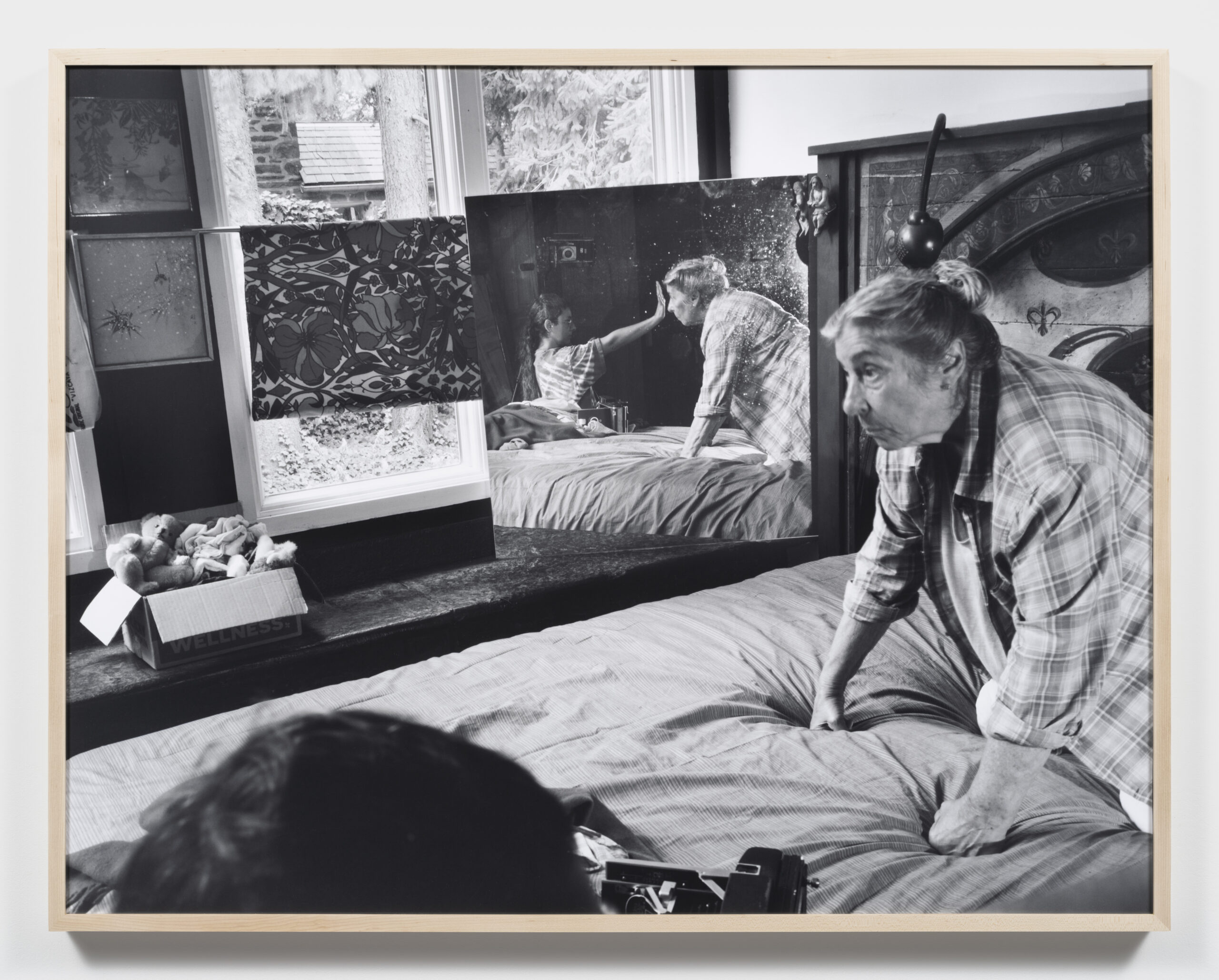 A black and white photograph depicts a woman sitting on a bed, looking intently at a reflection of a young girl in a mirror. The girl points toward the camera, and various objects, including a box of toys, are visible on the bed. Natural light filters through a window, illuminating the scene which conveys a sense of interaction and connection between the two figures.