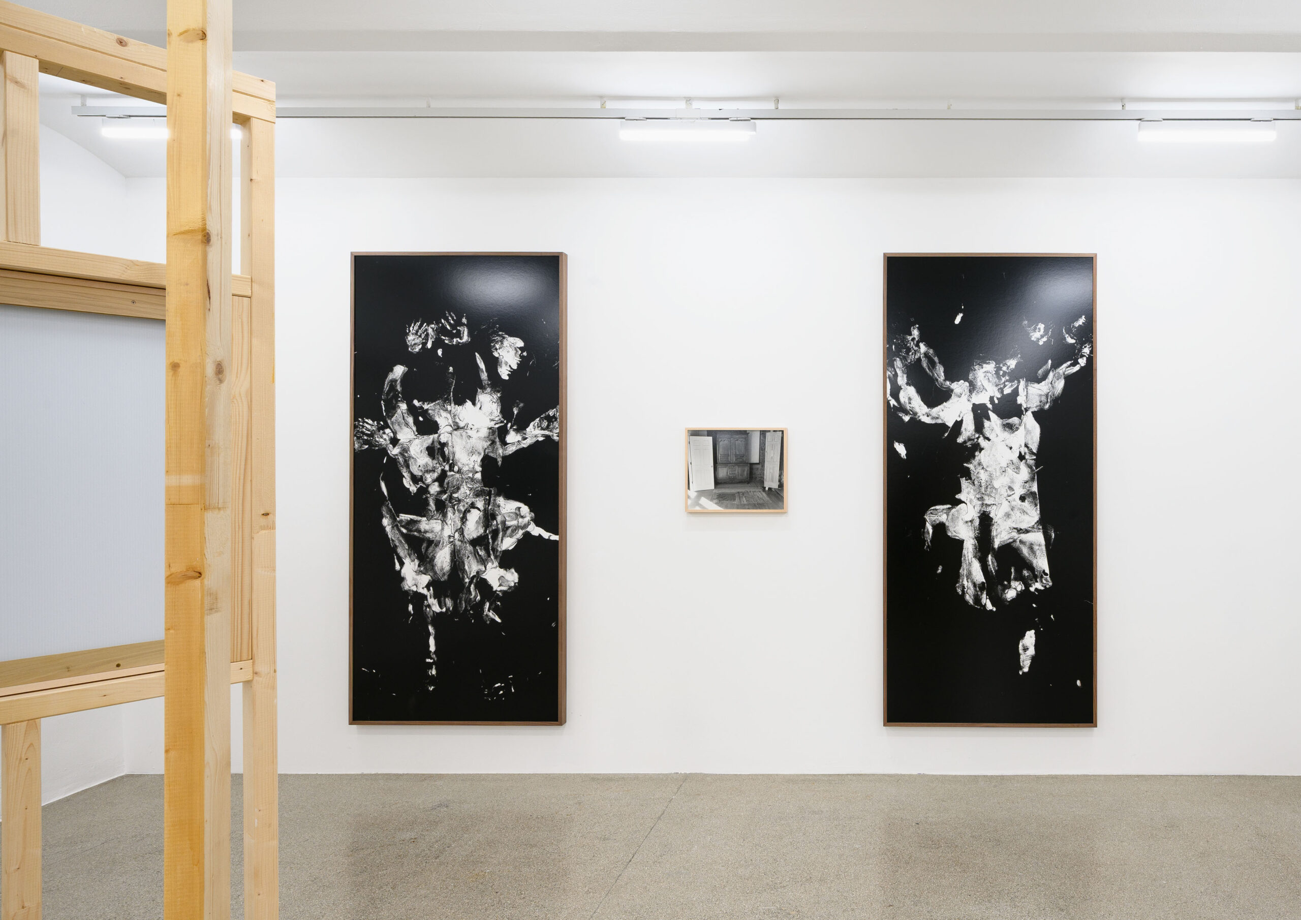 The image features an art gallery interior with three artworks on display. Two large black and white abstract pieces hang on the wall, depicting fluid forms and figures. In between them, a smaller framed artwork displays a black and white photograph of an interior space. The gallery space has a minimalist design, with wooden elements visible in the foreground and bright artificial lighting. The floor appears to be made of a smooth, polished material.