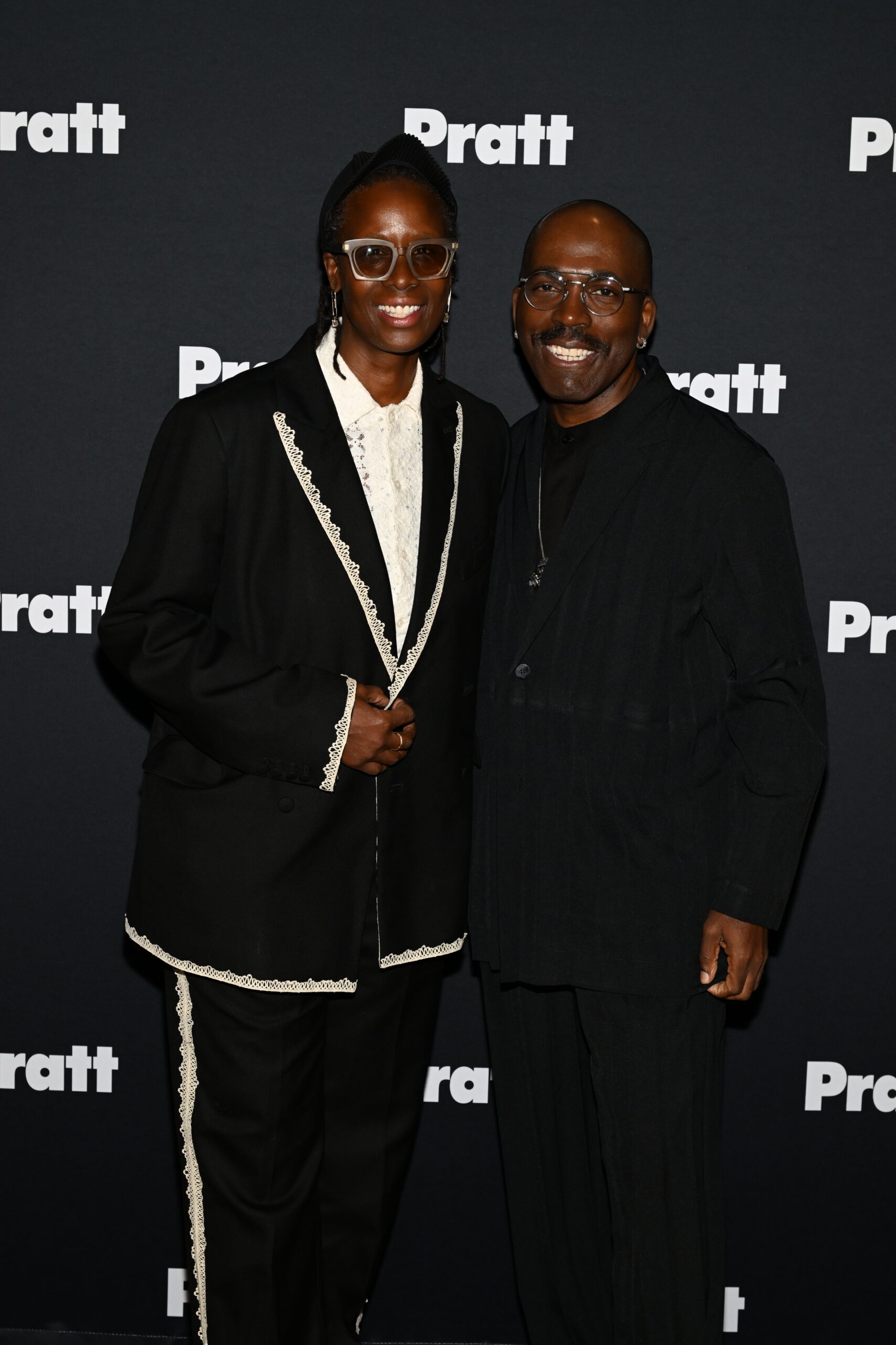 Two individuals are standing close together in front of a black backdrop featuring the word "Pratt" in white. One person is wearing a black suit with white lace detailing and large glasses, while the other is in a black outfit with a textured design. They are both smiling, exuding a friendly demeanor.