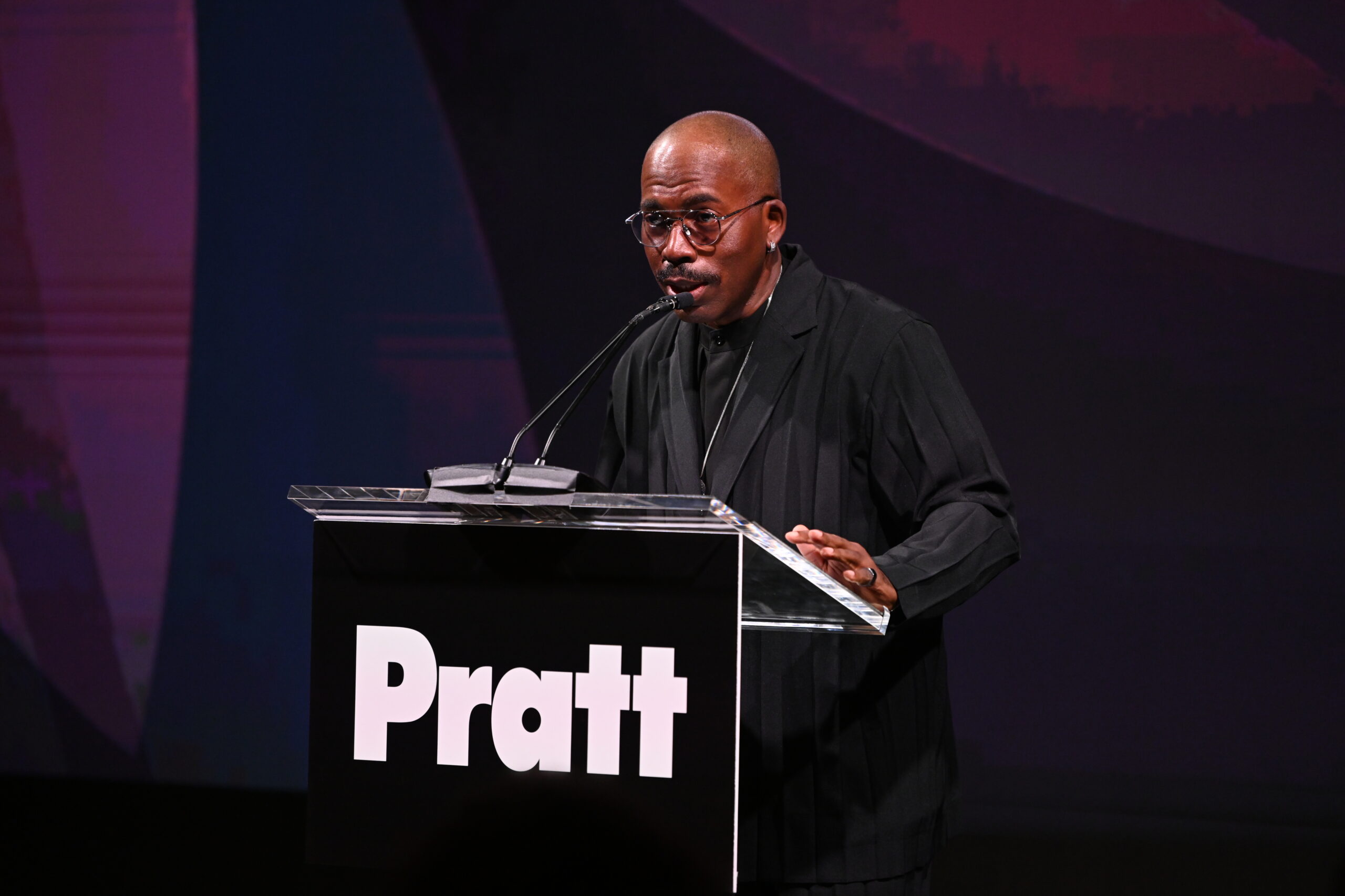A person with a shaved head and glasses speaks at a podium featuring the name "Pratt." They are dressed in a black, pinstriped outfit and are gesturing while addressing an audience. The background is slightly blurred with abstract patterns in muted colors, creating a dynamic atmosphere.