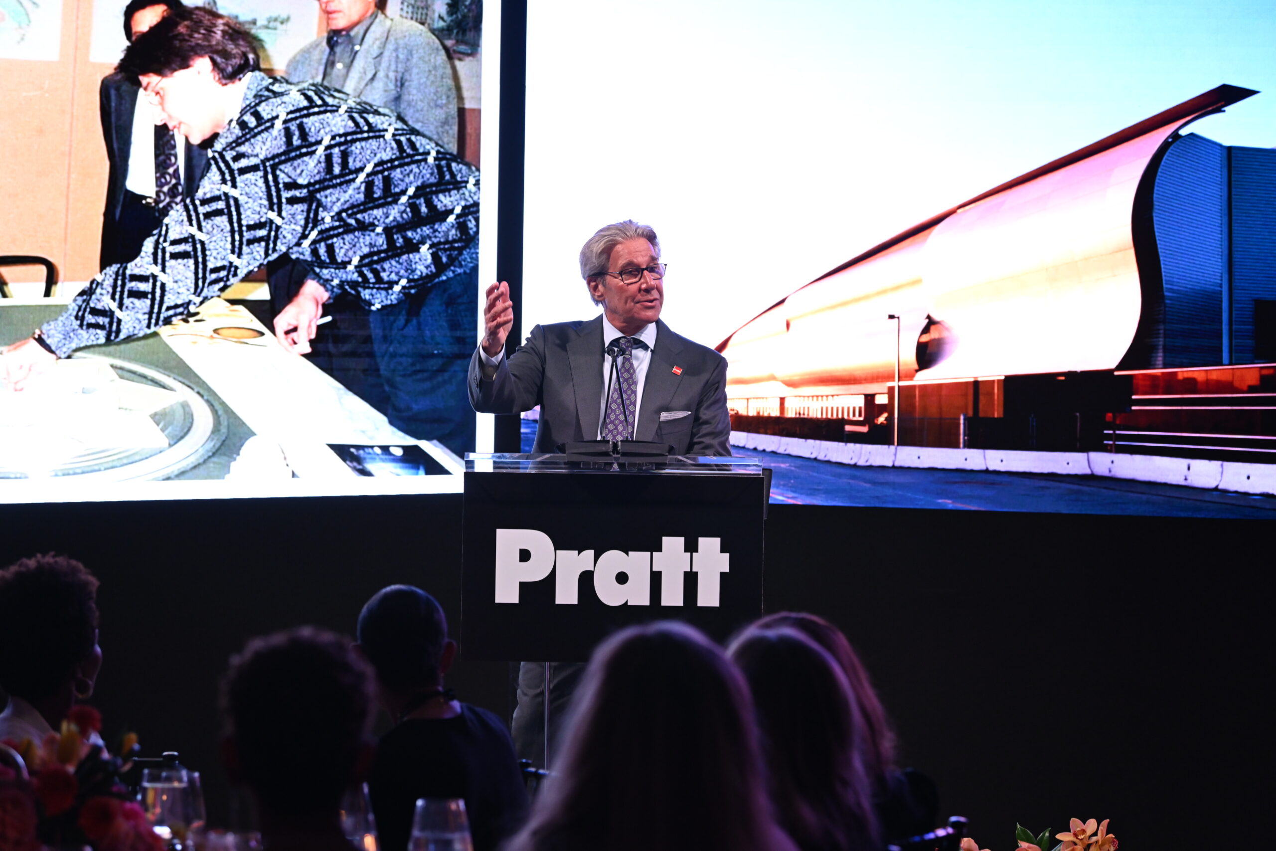 A speaker stands at a podium displaying the word "Pratt," addressing an audience at an event. Behind him is a large screen showing a historical image of a person working on a design. The audience's heads and shoulders are visible, and a floral arrangement is placed on the table in front of them.