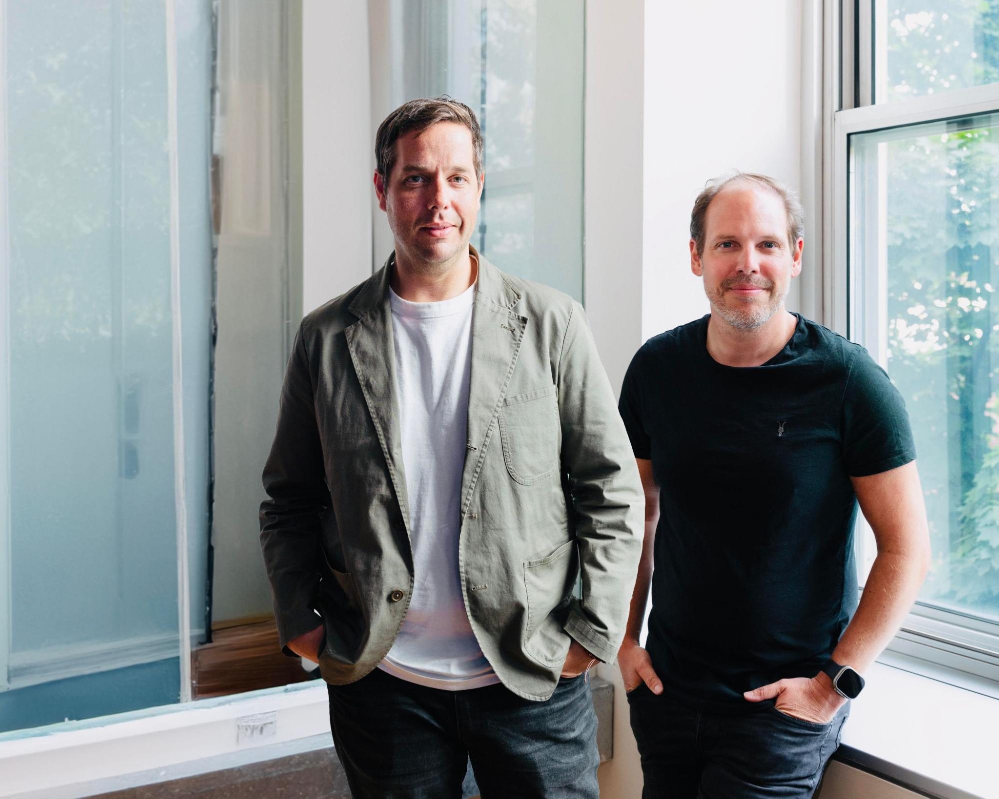 Two men standing side by side, both smiling slightly. The man on the left is wearing a light green jacket over a white t-shirt, while the man on the right is wearing a black t-shirt. They are indoors, with a large window to the right letting in natural light, and a frosted glass partition behind them. Both men have their hands in their pockets, with relaxed postures.