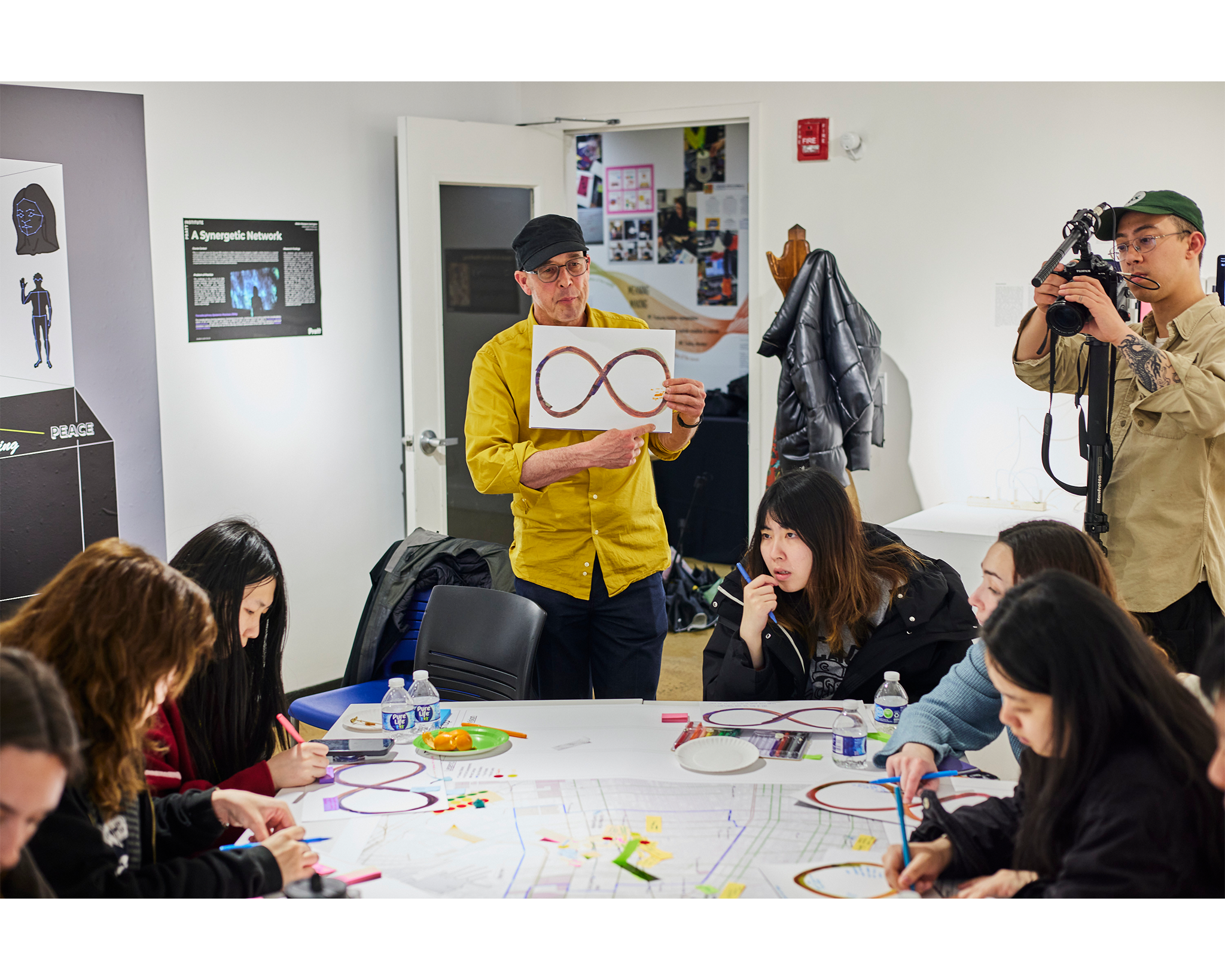 A man in a yellow shirt holds up a drawing of an infinity symbol while leading a classroom activity with students seated around a table, working on maps and drawings. A photographer on the right captures the moment as the students concentrate on their projects.