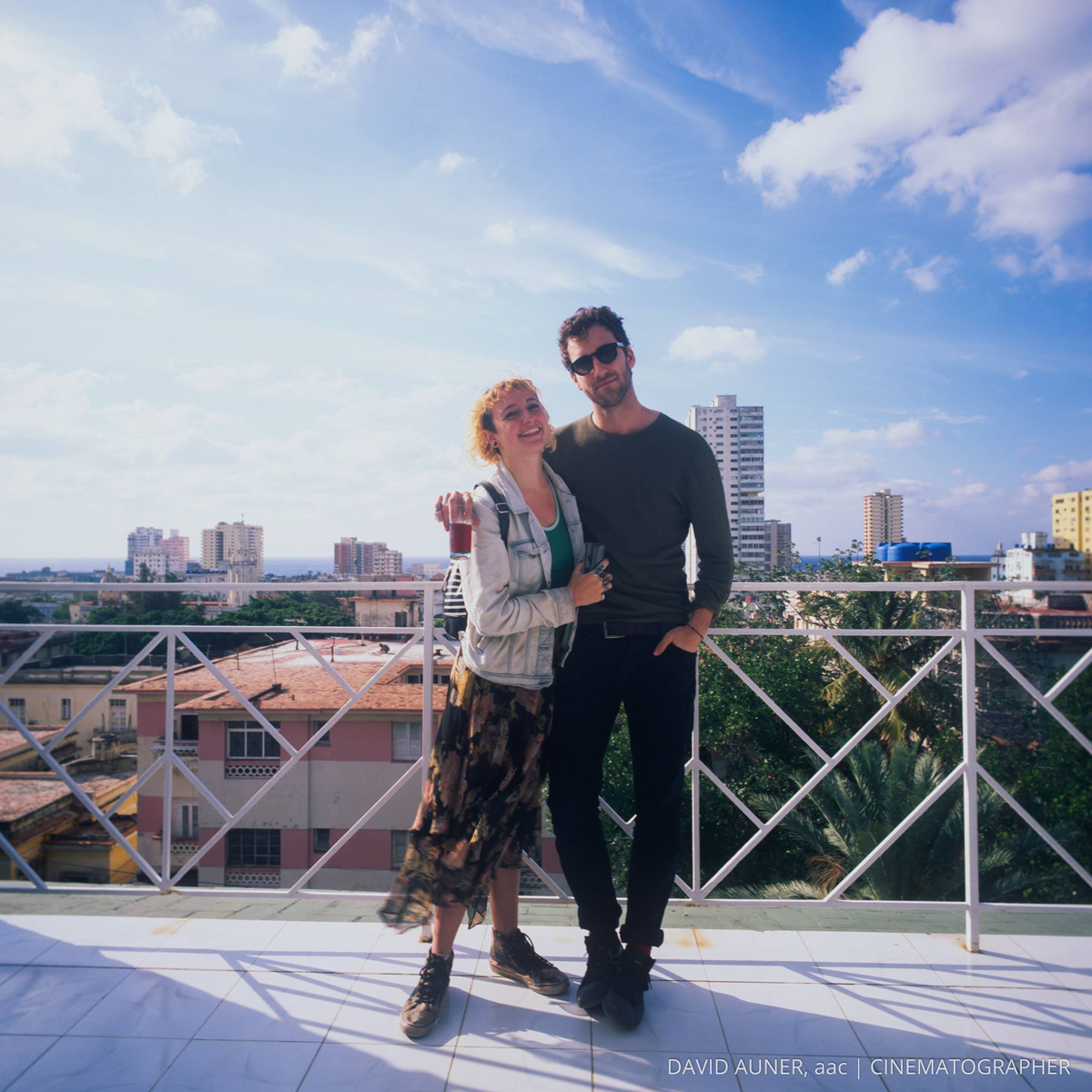 Kylie Shaffer and Charlie Cole standing together on a rooftop balcony with a big blue sky and buildings and trees behind them.