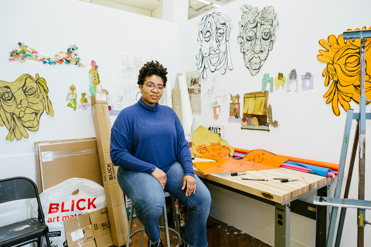 Devin Alexander sitting on a stool in her studio surrounded by her artwork.