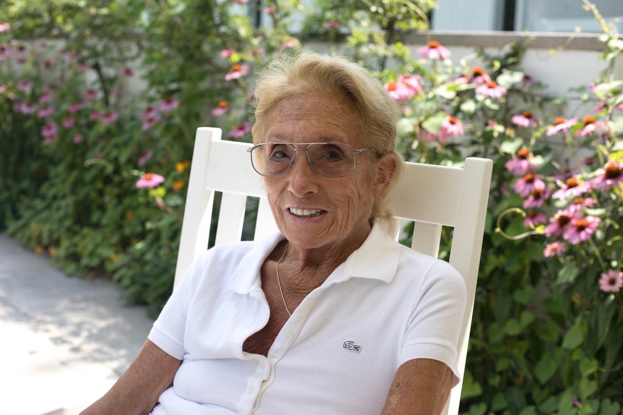 A photo of Hazel Siegel sitting in a white chair outside in a garden with flowers behind her.