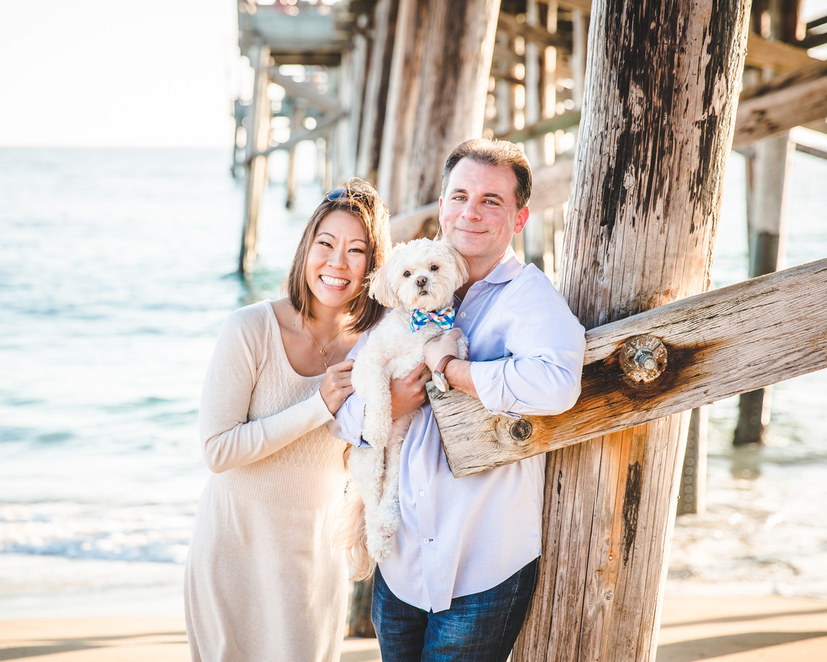 Cynthia Kwan Knotz and Christopher Knotz standing by a pier smiling. Christopher is holding their small white dog.