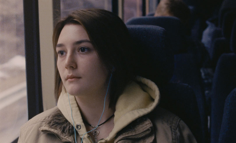 Still from Never Rarely Sometimes Always. A young woman sitting on a bus looks out the window. She is wearing a tan winter coat over a yellow sweatshirt. She has her earbuds in.